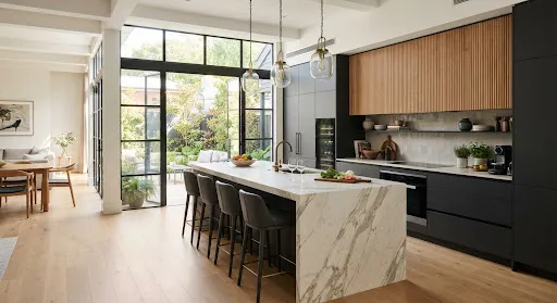 High-end open plan designer kitchen with massive Calacatta gold waterfall marble island, sleek charcoal matte cabinets, fluted natural timber upper cabinets, pendant glass lighting, wine fridge, floor-to-ceiling black steel casement windows with courtyard garden view.
